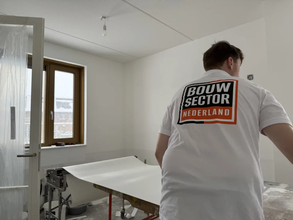 Behanger van Bouwsector Nederland trekt een baan renovlies over de plaktafel in een kamer met houten kozijn